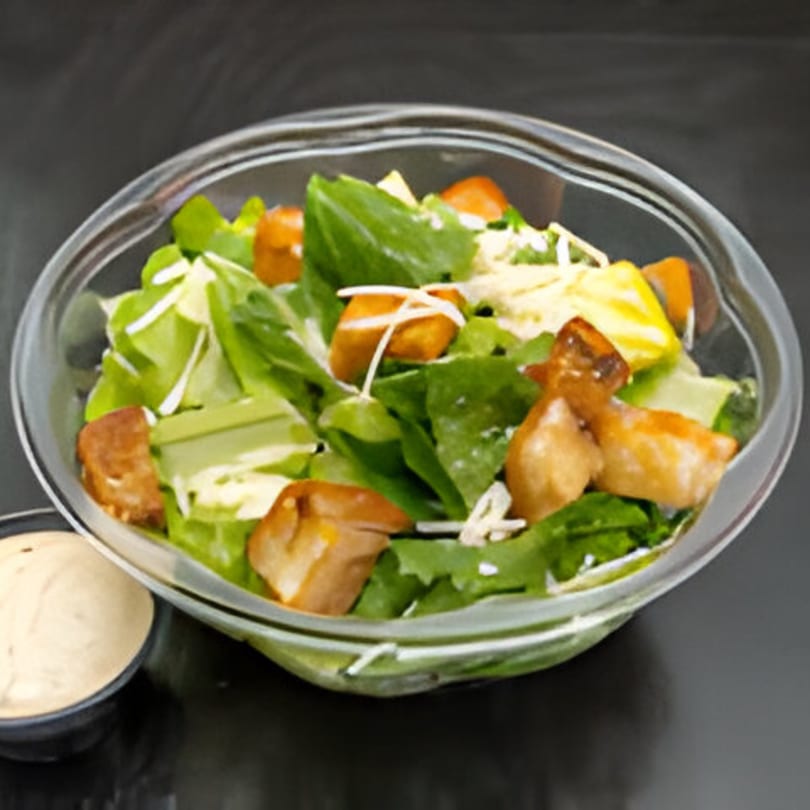 A fresh green salad with croutons and shredded cheese in a clear bowl, with dressing on the side.
